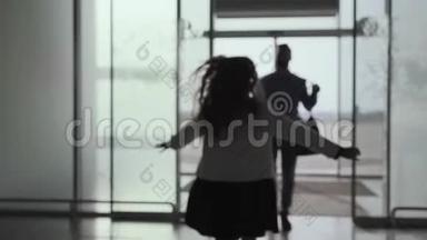 白种人年轻人带着行李走进玻璃机场门的剪影。 一个快乐的小女孩跑过来